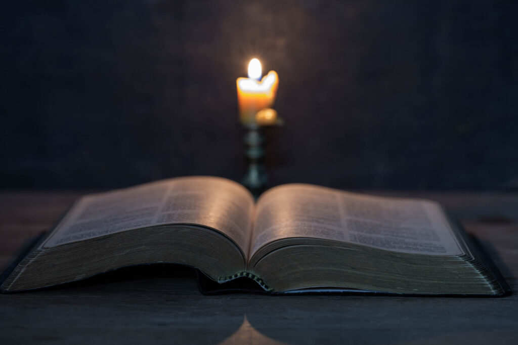 An open book rests on a wooden surface, illuminated by the warm glow of a candle in the background, creating a serene reading atmosphere.