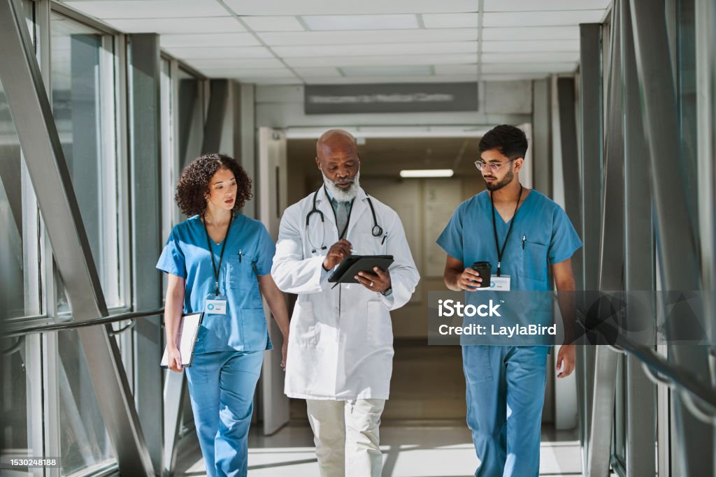 Three healthcare professionals in scrubs walk through a hospital corridor. One doctor reviews a tablet, while the others engage in conversation.